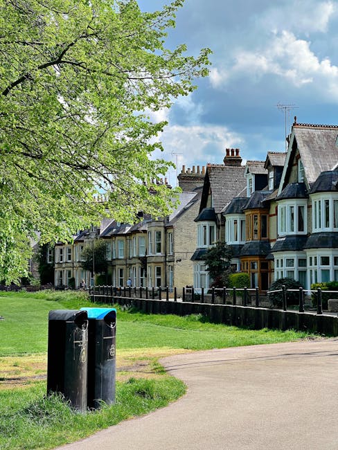 A row of traditional terraced houses with bay windows and slate roofs situated along a residential street, partially obscured by lush green trees with fresh spring foliage. In the foreground, two black wheelie bins are positioned on a paved pathway adjacent to a small grassy area, which is bordered by a low wooden fence separating the pavement from the front gardens of the houses. The sky above is partly cloudy with patches of blue, indicating a clear day. The scene reflects a typical urban or suburban environment where private rubbish collection might be managed independently, aligned with services like waste collection Balham. The layout and setting suggest a peaceful neighborhood with on-site waste management options that support local rubbish clearance activities, complementing broader waste disposal practices in the area.