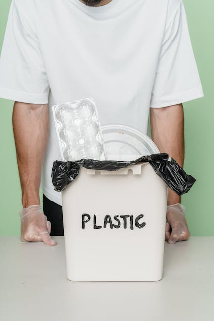 A person wearing a white T-shirt and transparent disposable gloves stands behind a white plastic bin labeled 'PLASTIC' in black text. The bin is partly covered with a black garbage bag, which is folded over the top edge. Inside the bin, there is an empty transparent plastic blister pack with three circular cavities visible. The background features a plain, light green wall, and the scene is evenly lit, emphasizing the focus on the plastic waste segregation process. This setup reflects an emphasis on proper plastic recycling practices, which are often part of independent waste collection or rubbish removal services. The image underscores the importance of correctly sorting recyclable materials for efficient disposal or recycling, aligning with the broader context of rubbish and waste management solutions provided by companies like wastecollectionbalham.co.uk.