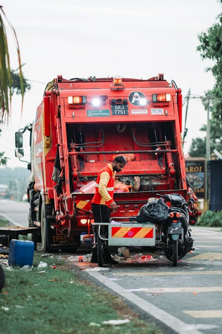 A worker wearing a yellow safety vest and black pants is operating a red rubbish collection vehicle on a street. The vehicle has its rear compactor open, revealing a metal drum and mechanical components used for rubbish compression, with some waste visible at the base. The worker is standing on the pavement, leaning slightly forward, with a black plastic bag placed on a metal platform attached to the vehicle. Nearby, a small black motorcycle is parked alongside a white line marking the edge of the road, with additional items such as a blue container and some scattered debris visible on the ground. In the background, there are trees, electrical wires, and a commercial building with signage, indicating an urban environment. The scene is lit by natural daylight, and the setting suggests independent waste collection or on-site clearance as part of private rubbish removal services, with emphasis on operational activities involving waste management equipment.
