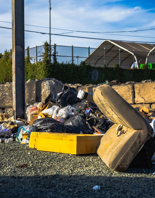A worker wearing a yellow safety vest and black pants is operating a red rubbish collection vehicle on a street. The vehicle has its rear compactor open, revealing a metal drum and mechanical components used for rubbish compression, with some waste visible at the base. The worker is standing on the pavement, leaning slightly forward, with a black plastic bag placed on a metal platform attached to the vehicle. Nearby, a small black motorcycle is parked alongside a white line marking the edge of the road, with additional items such as a blue container and some scattered debris visible on the ground. In the background, there are trees, electrical wires, and a commercial building with signage, indicating an urban environment. The scene is lit by natural daylight, and the setting suggests independent waste collection or on-site clearance as part of private rubbish removal services, with emphasis on operational activities involving waste management equipment.